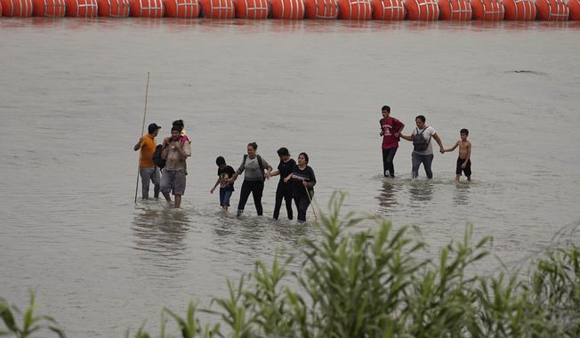 Migrants who crossed the Rio Grande from Mexico walk past large buoys being deployed as a border barrier on the river in Eagle Pass, Texas, Wednesday, July 12, 2023. The floating barrier is being deployed in an effort to block migrants from entering Texas from Mexico. (AP Photo/Eric Gay)