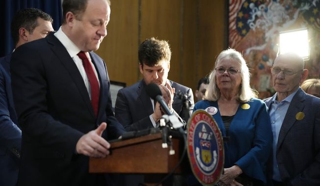 FILE - Colorado Gov. Jared Polis, front left, pauses as he speaks as Sandy Phillips, second from right, and her husband, Lonnie, who lost their daughter in the mass shooting at a theatre in Aurora, Colo., look on before Polis signed four gun control bills into law during a ceremony April 28, 2023, in the State Capitol in Denver. A federal judge on Monday, Aug. 7, has blocked Colorado from enforcing a new law raising the age to purchase any gun from 18 to 21. (AP Photo/David Zalubowski, File)