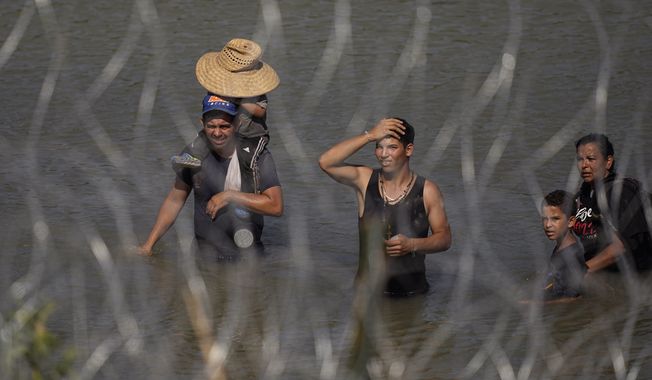 Migrants stand in the Rio Grande behind concertina wire as they try to enter the U.S. from Mexico near the site where workers are assembling large buoys to be used as a border barrier in Eagle Pass, Texas, Tuesday, July 11, 2023. The floating barrier is being deployed in an effort to block migrants from entering Texas from Mexico. (AP Photo/Eric Gay)